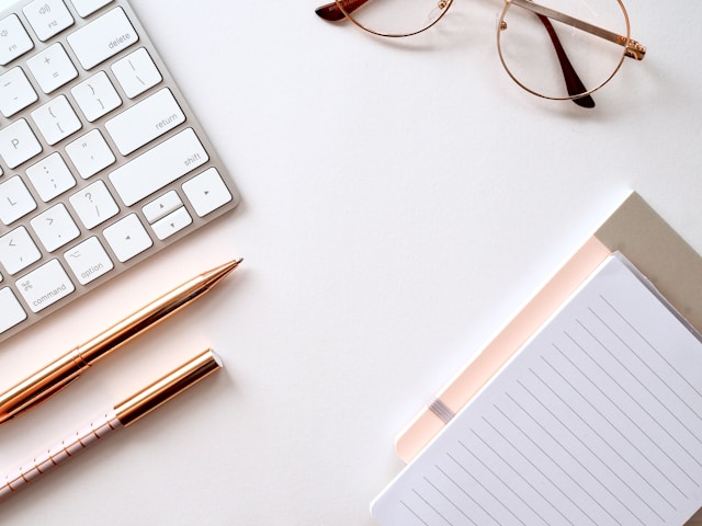 picture of a flatlay with a keyboard, pens, glasses and a notebook